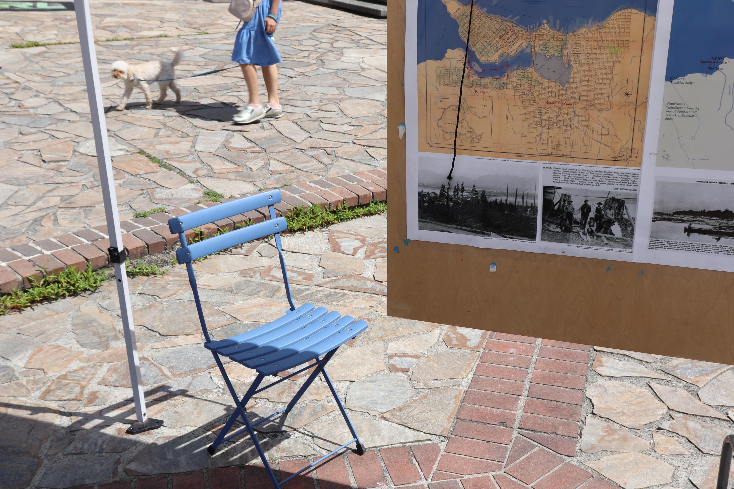 An empty blue folding bistro chair on a flagstone plaza, a dog-walker passing behind it and a historical map display to the right.