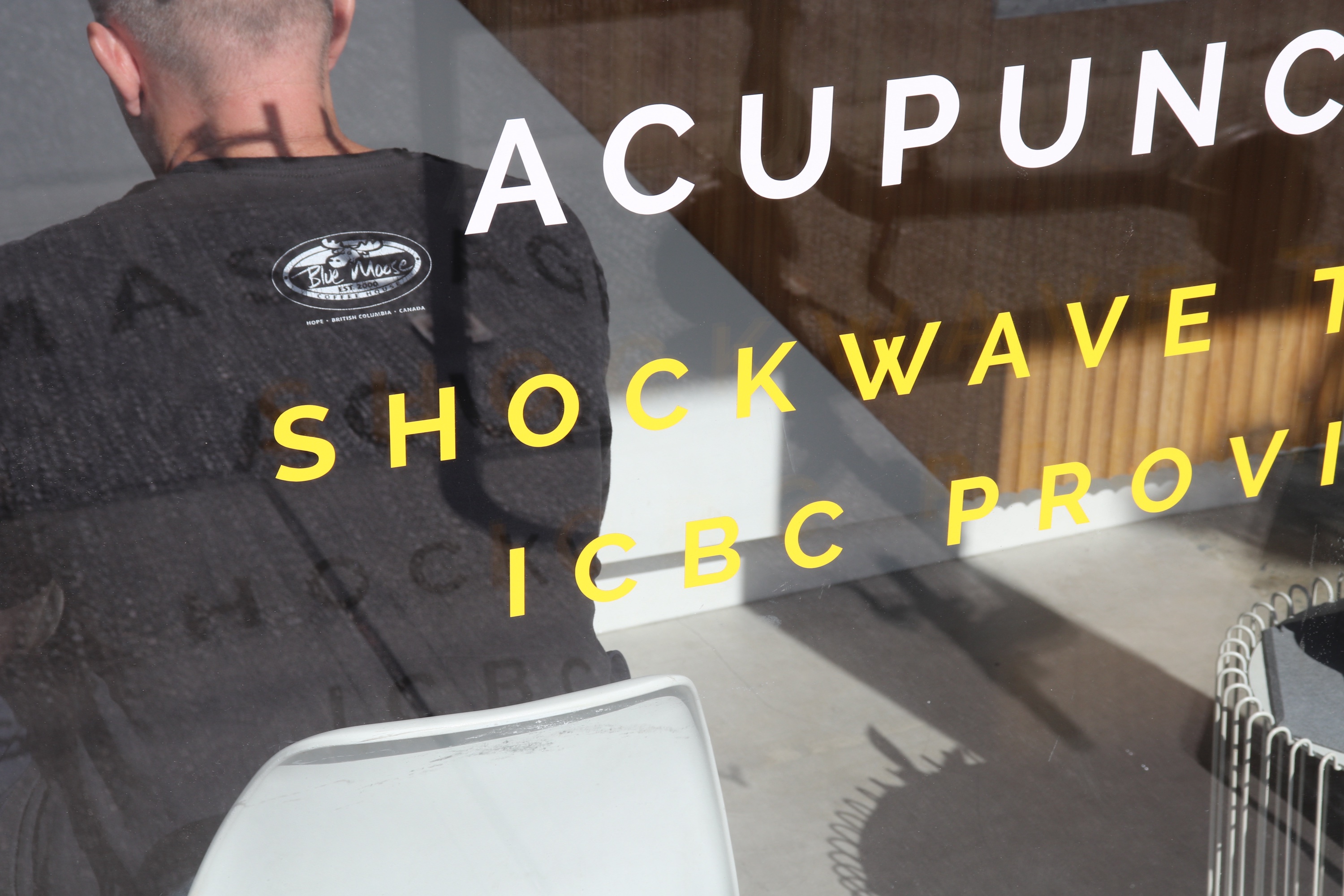 Seen through a clinic window: a man in a Blue Moose sweatshirt from behind, overlaid with the words ACUPUNCTURE and SHOCKWAVE.