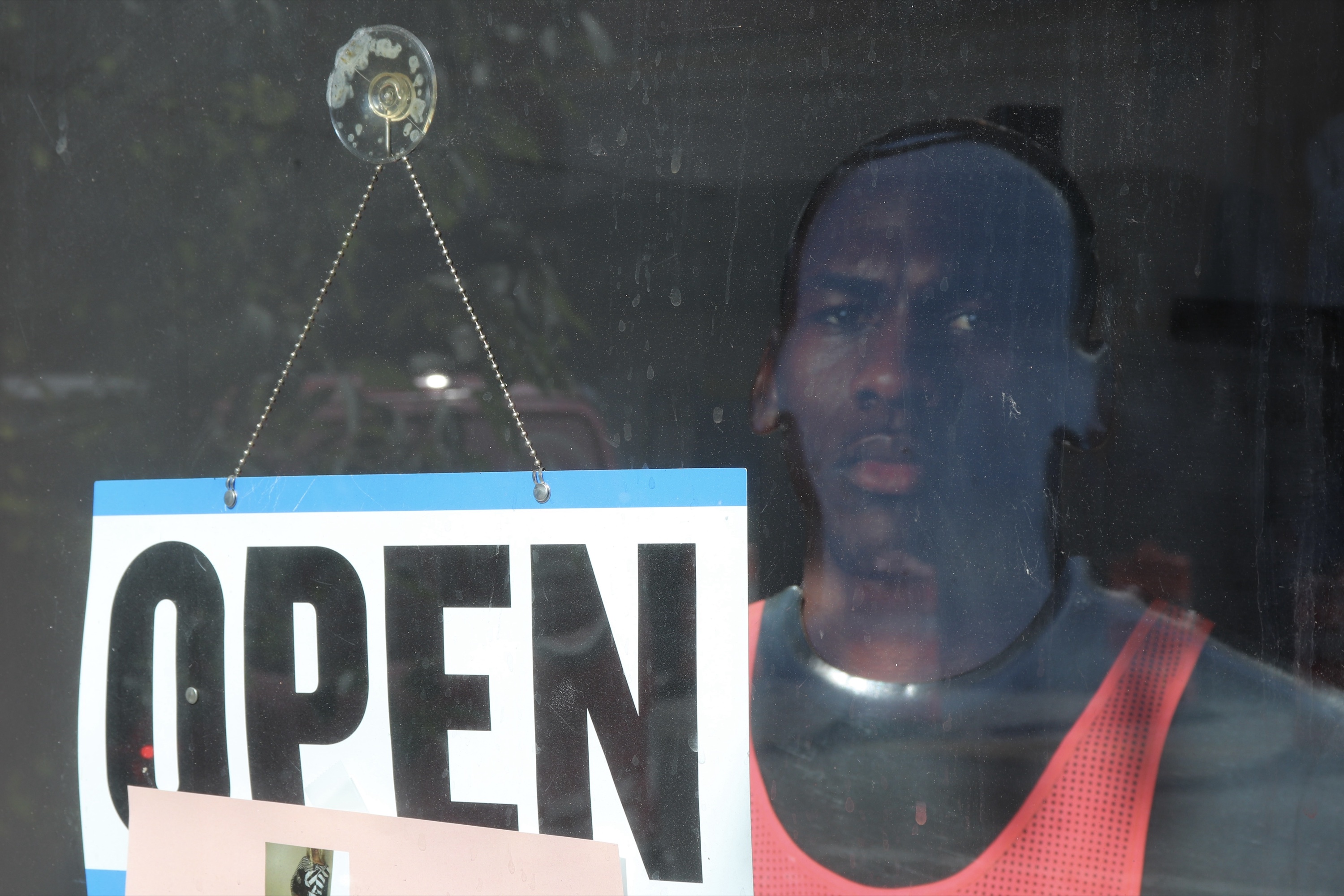 Shot from the sidewalk through a shop window: a hanging OPEN sign, and inside a mannequin of a man in a red mesh tank top gazing out.