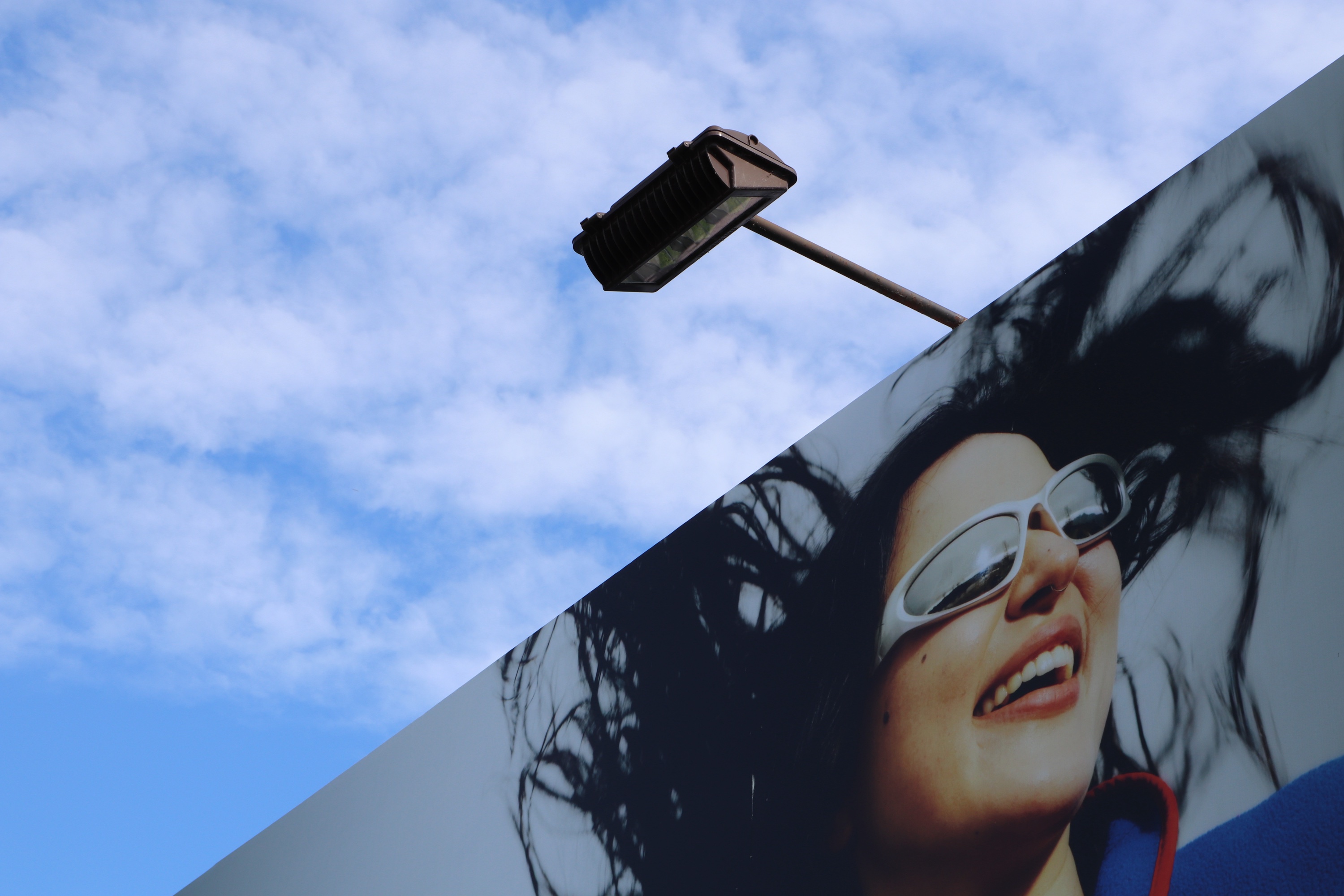 Looking up at a billboard of a smiling woman in white sunglasses, with a floodlight arm against a cloud-streaked blue sky.
