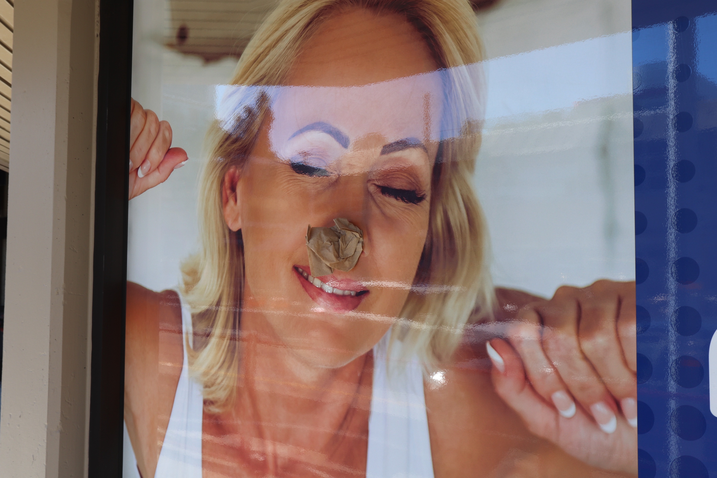 A sun-worn advertising poster behind glass showing a blonde woman with closed eyes, partly stripped and patched.