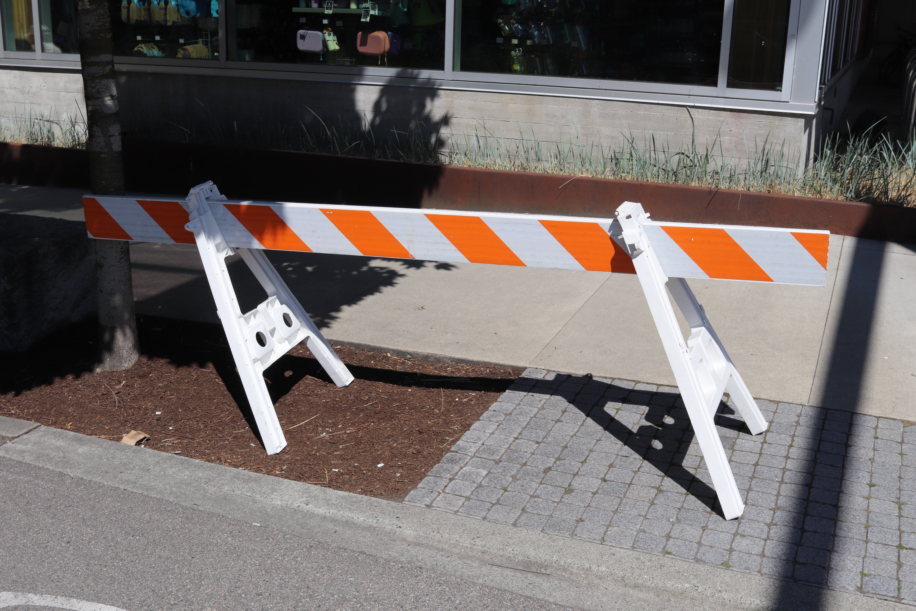 A white plastic road barricade leaning to one side, its orange-and-white striped rail in bright sunlight outside a shop window.