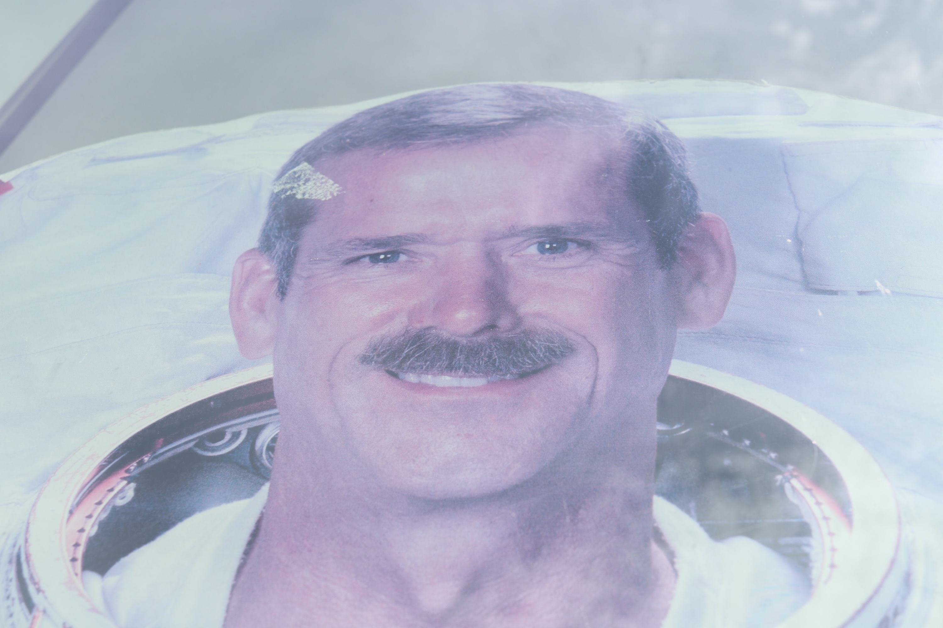 A faded pink-and-blue printed portrait of a smiling moustached astronaut, closely framed behind a reflective surface.