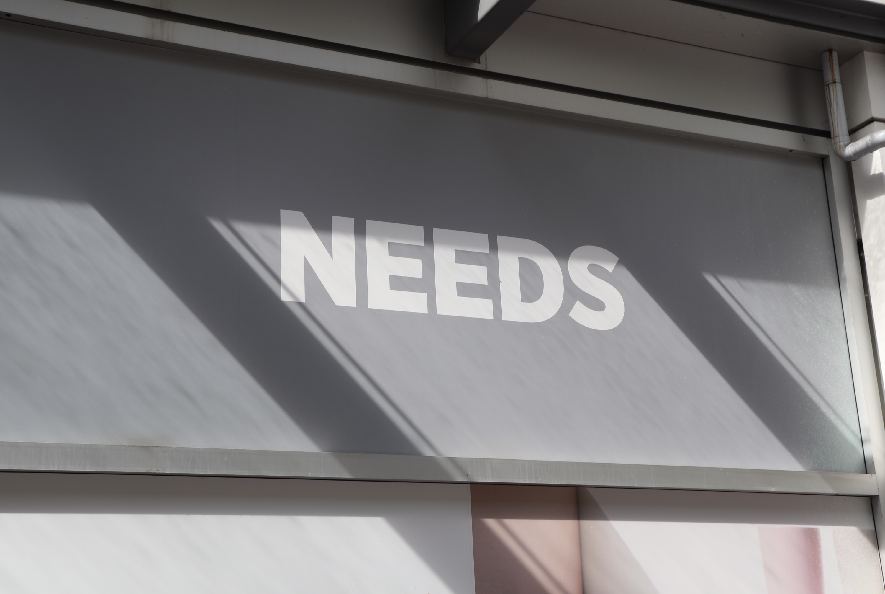 The word NEEDS in white capital letters on a grey storefront panel crossed by sharp diagonal shadows.