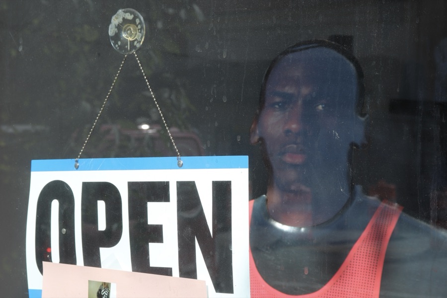 Shot from the sidewalk through a shop window: a hanging OPEN sign, and inside a mannequin of a man in a red mesh tank top gazing out.