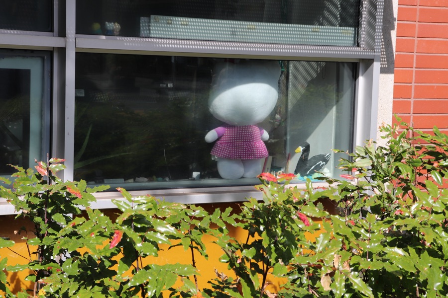 A white soft-toy figure in a pink polka-dot dress standing on a windowsill above a hedge of oak-leaf foliage.