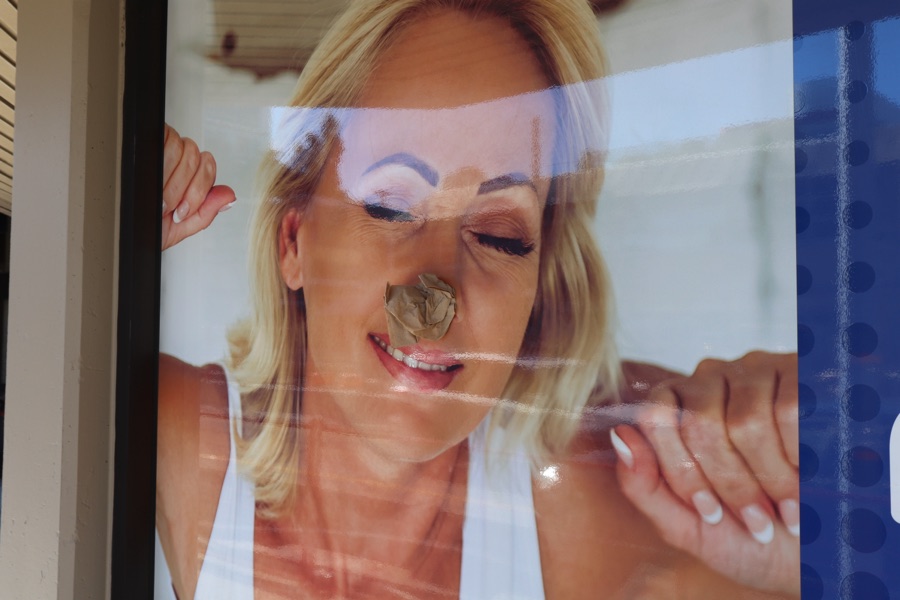 A sun-worn advertising poster behind glass showing a blonde woman with closed eyes, partly stripped and patched.