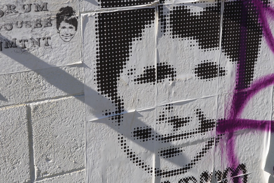 Large and small versions of the same smiling face pasted on a masonry wall, with the halftone portrait crossed by purple spray paint and hard shadows.