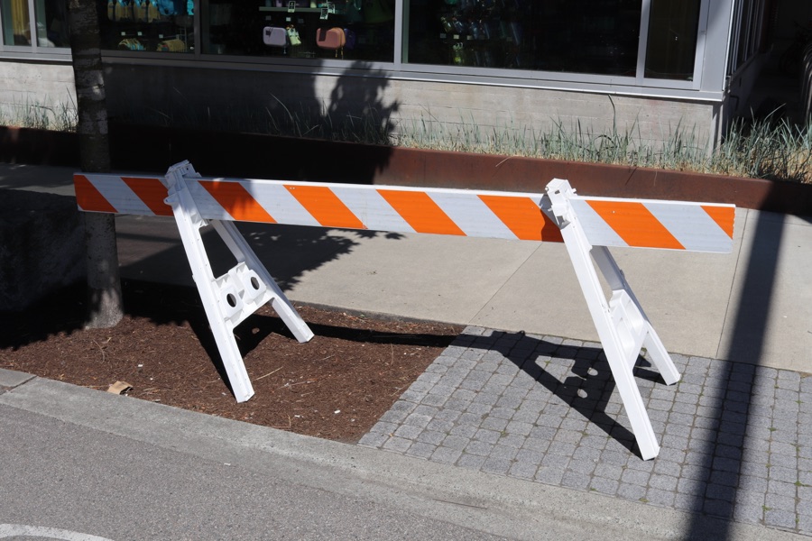 A white plastic road barricade leaning to one side, its orange-and-white striped rail in bright sunlight outside a shop window.