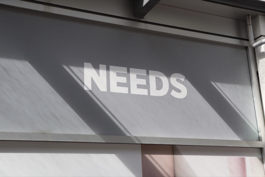 The word NEEDS in white capital letters on a grey storefront panel crossed by sharp diagonal shadows.